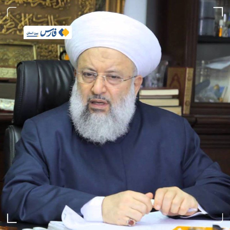 A bearded man wearing a white turban and dark robe sits at a desk with papers, speaking in a room featuring Arabic calligraphy.