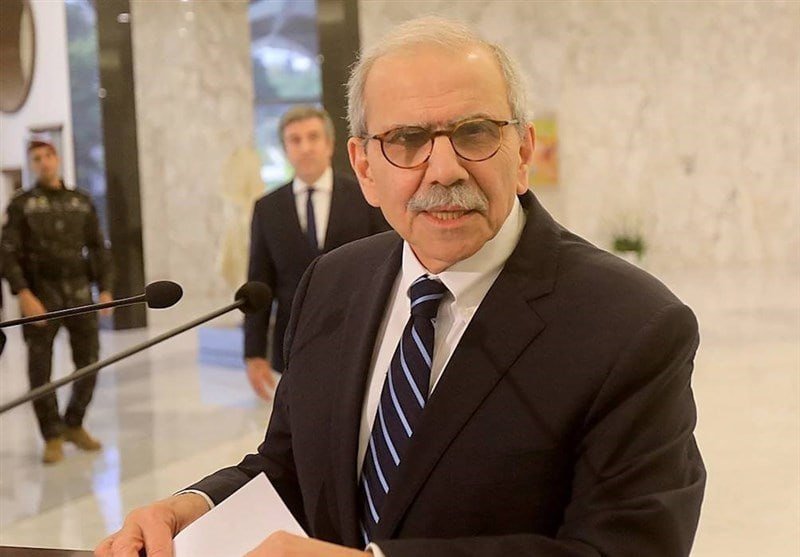 An older man with glasses, a mustache, and a striped tie stands at a podium with microphones, holding a paper, while another suited man and a uniformed guard stand behind him.