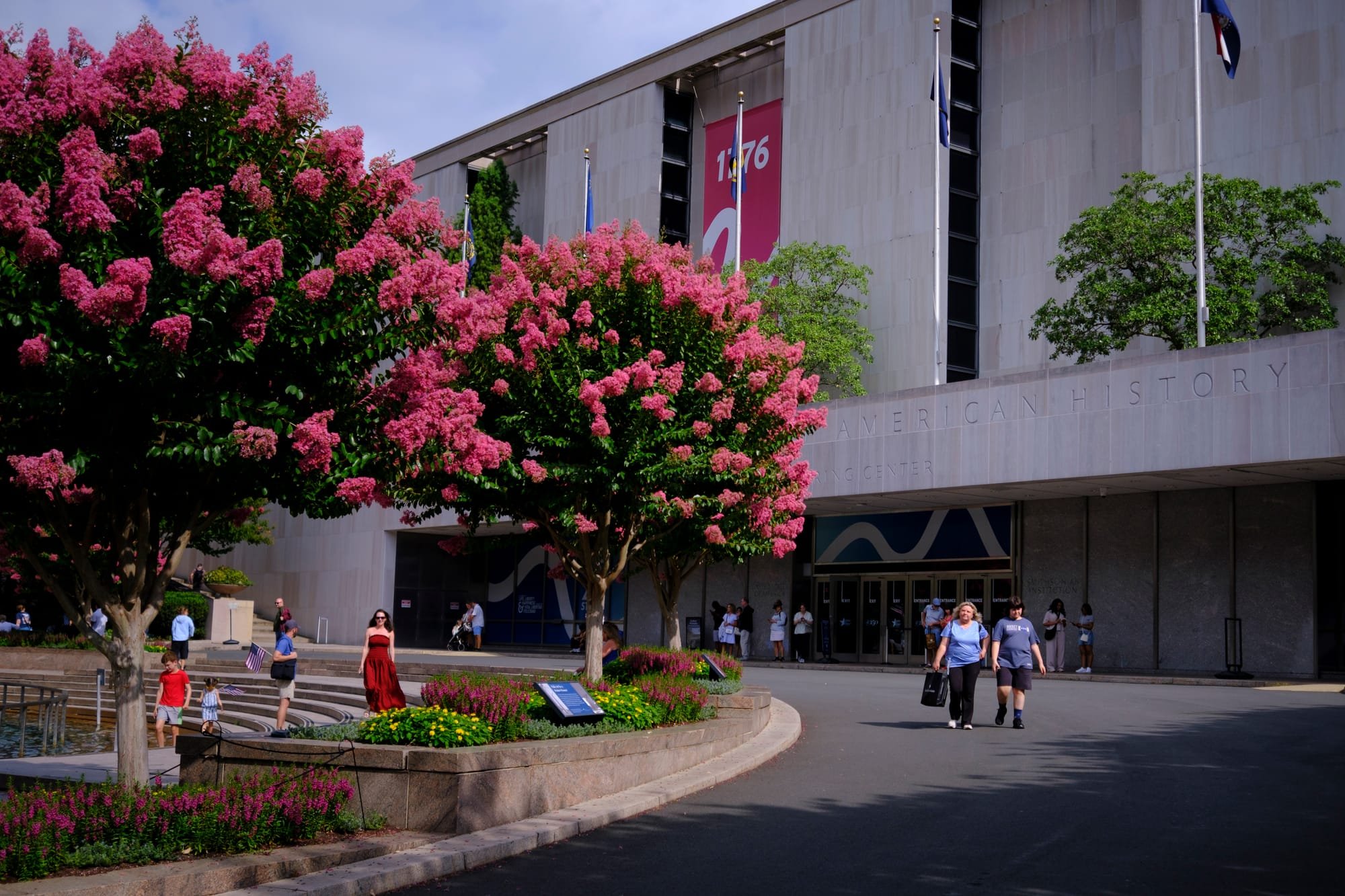 Visitors walk past blooming pink crepe myrtle trees outside the entrance of the American History museum, where a 1776 banner hangs on the facade.