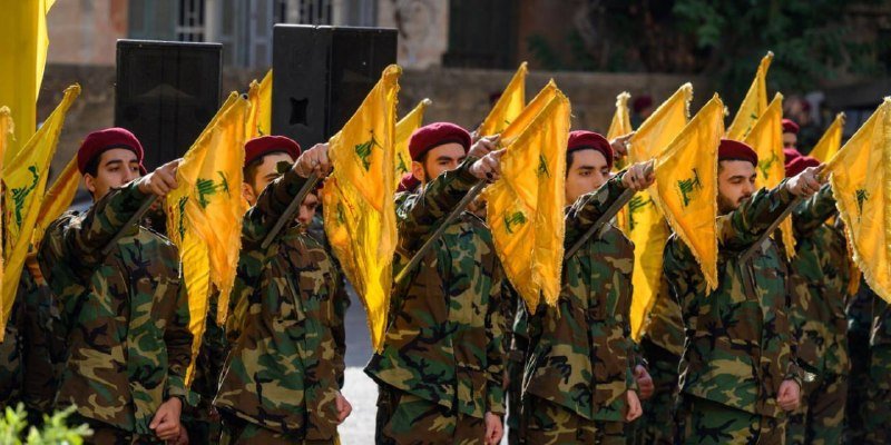 Soldiers in camouflage uniforms and maroon berets march in formation while carrying yellow flags featuring a green emblem, with large black speakers visible in the background.