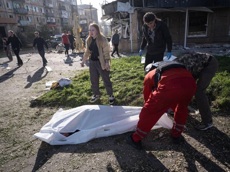 Emergency responders in red cover a body bag on grass amid debris and damaged buildings while bystanders watch in an urban setting.