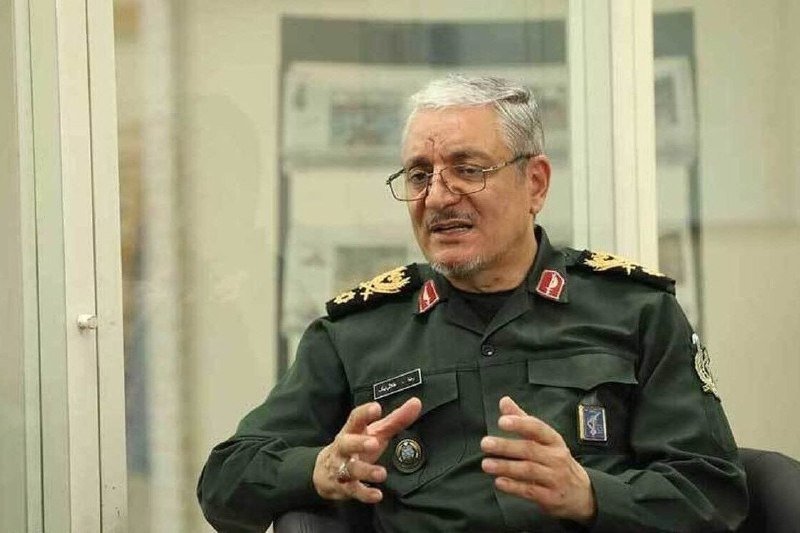 A gray-haired man with glasses, wearing a green military uniform with insignia, gestures with both hands while speaking in an indoor setting.