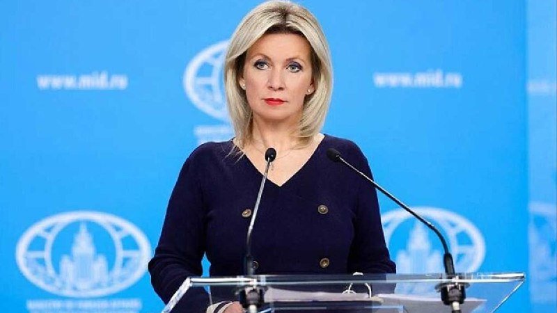 A blonde woman in a dark blue jacket stands at a clear podium with microphones, speaking in front of a blue backdrop displaying "www.mid.ru" and a globe logo.