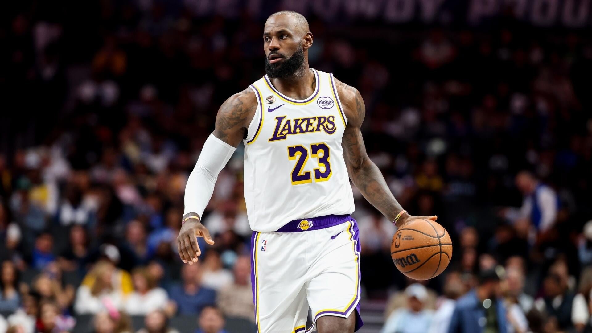 A bearded basketball player wearing a white Lakers jersey with the number 23 dribbles a basketball during a game, with a crowd visible in the background.