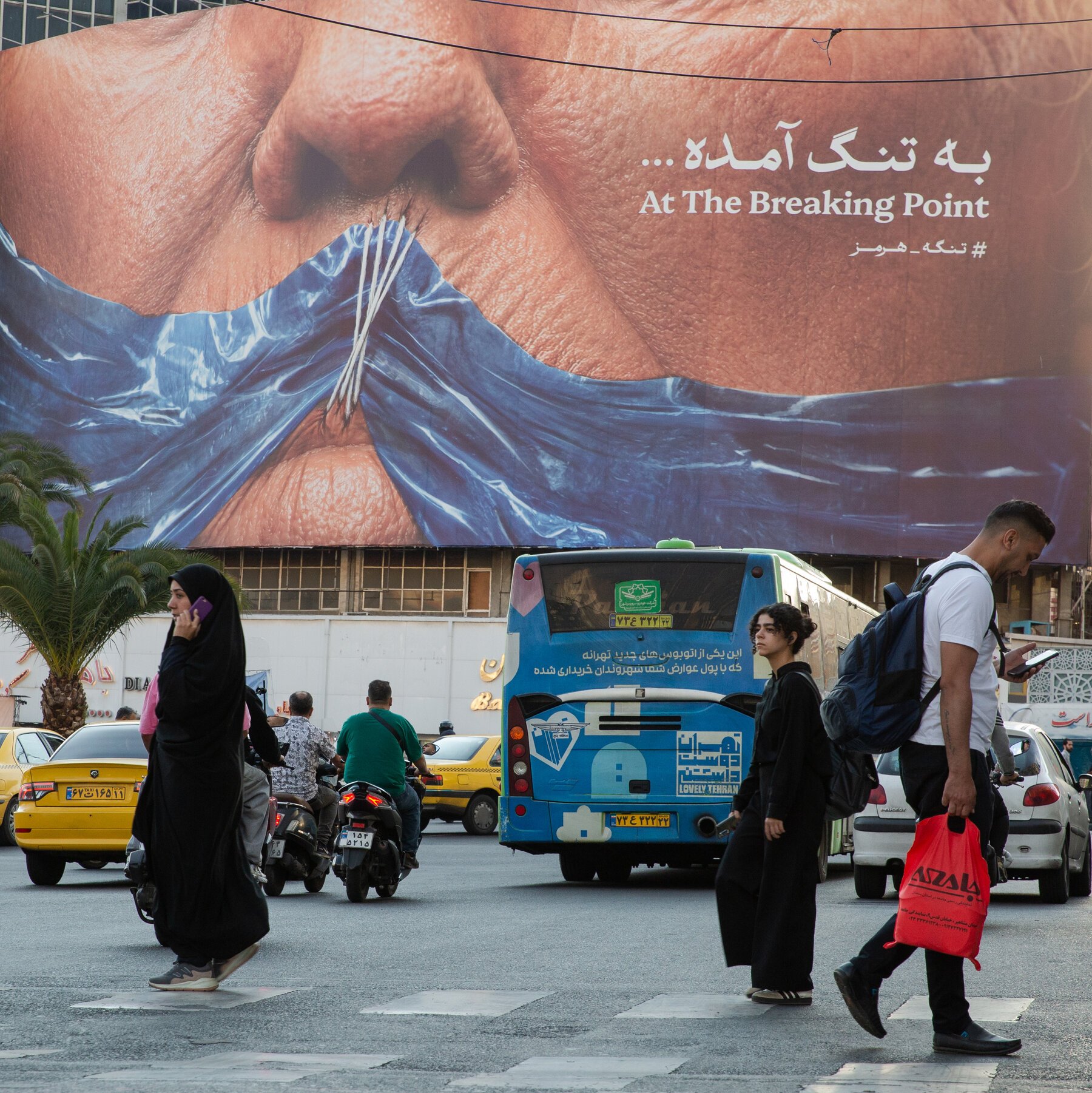 Pedestrians cross a busy street as taxis, motorcycles, and a blue bus pass in front of a massive billboard displaying a close-up of a stitched surgical wound with the text "At The Breaking Point."