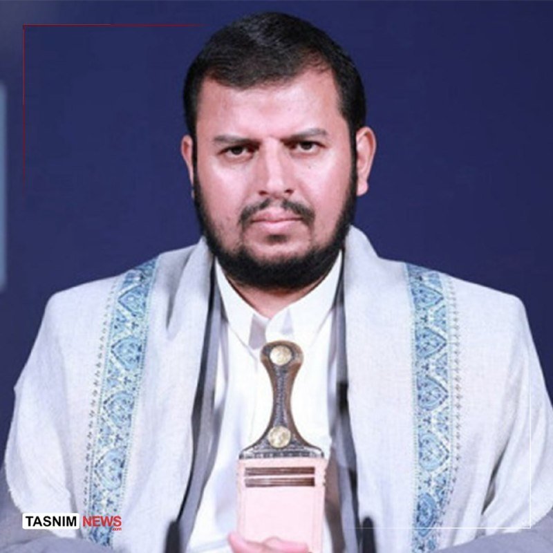 A bearded man in a light gray patterned jacket holds a decorative trophy with a curved handle, posed against a dark blue background, with a "Tasnim News" logo visible.