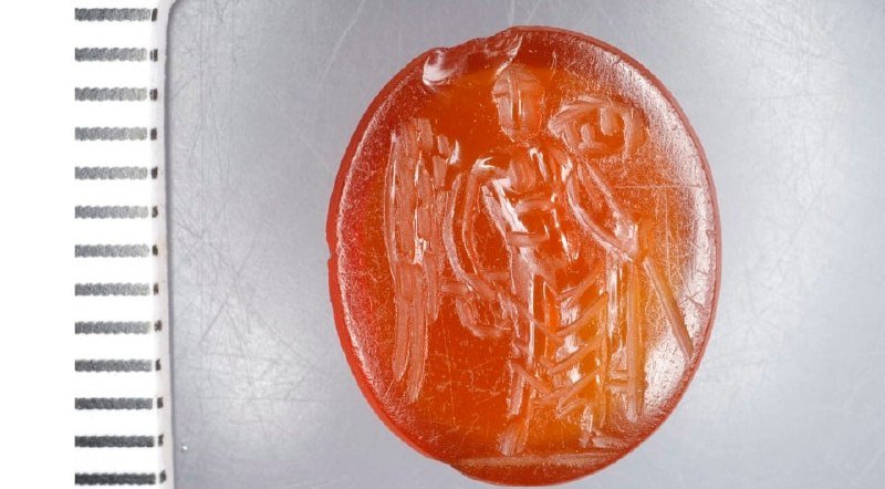 A red wax seal depicting a carved figure of a knight or soldier stands beside a spiral-bound notebook on a scratched metallic surface.