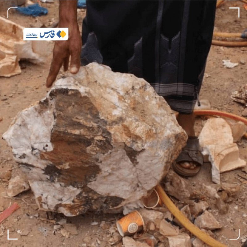 A person wearing a dark robe and sandals holds a large piece of crystalline rock on a debris-strewn, sandy surface scattered with stone fragments and hoses.