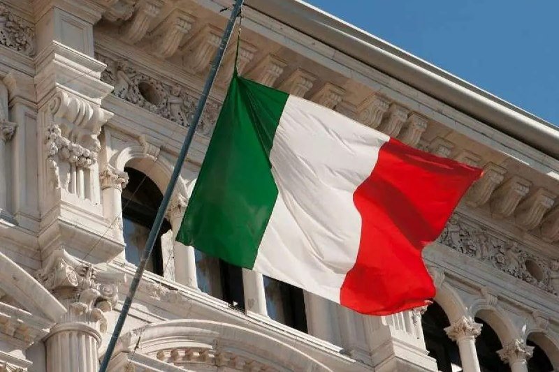 An Italian flag waves in front of an ornate, light-colored building facade with decorative columns and architectural details under a clear sky.