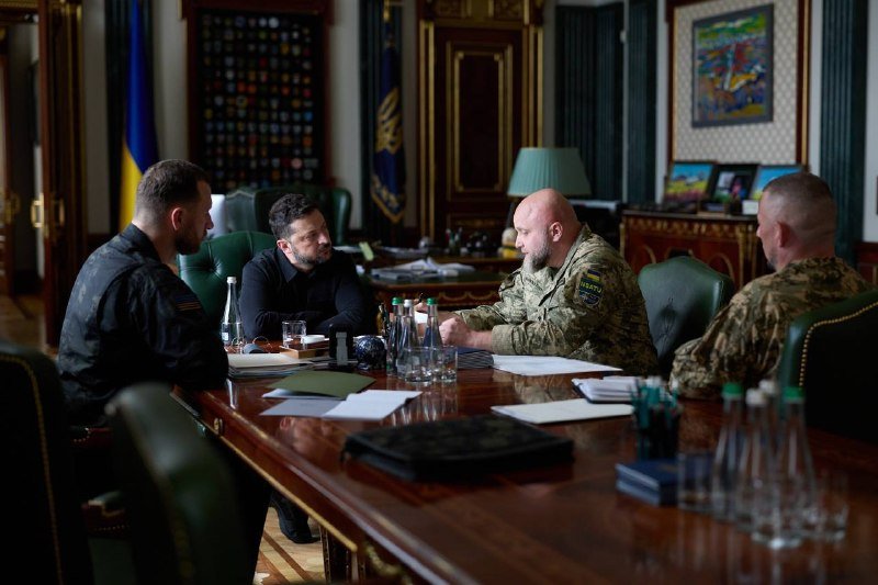 Four men, including one in a dark sweater and others in military uniforms, sit across a polished conference table during a meeting in an ornate room with a Ukrainian flag.