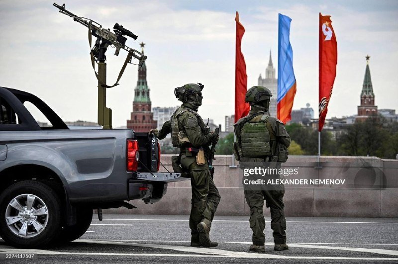 Two armed soldiers in green uniforms stand beside a mounted machine gun on a pickup truck, with Kremlin towers and red and blue flags visible in the background.