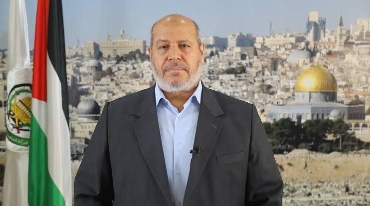 A gray-bearded man in a dark suit and light blue shirt speaks indoors, with a Palestinian flag visible on the left and the Dome of the Rock and Jerusalem skyline in the background.