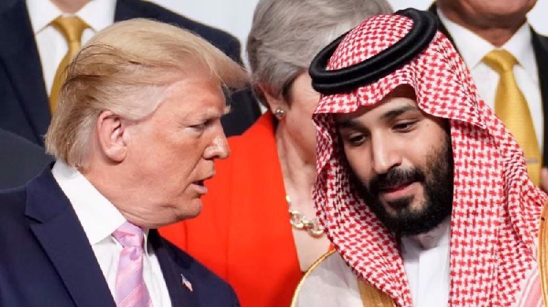 Two men in formal attire—one in a dark suit with pink tie, the other in a traditional red-and-white checkered headdress—stand near each other, with other formally dressed individuals in the background.