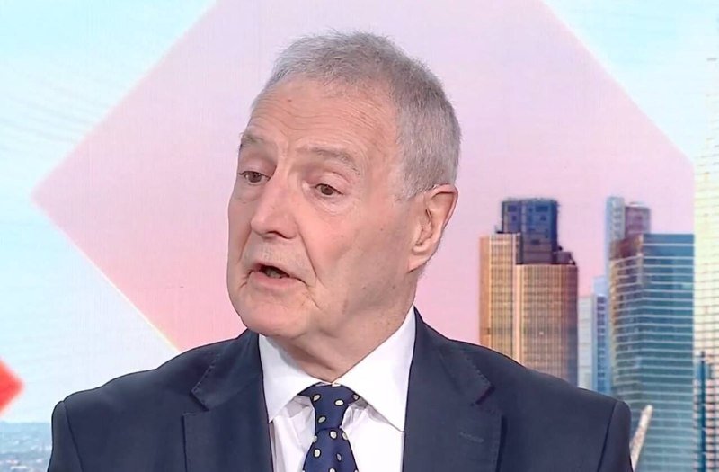 An older man with gray hair wearing a dark suit, white shirt, and dotted tie speaks in front of a studio backdrop featuring a city skyline and geometric shapes.