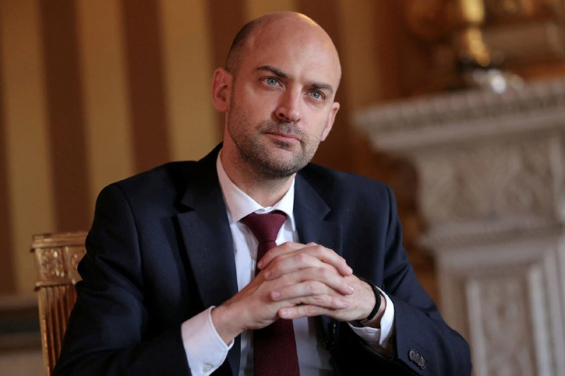A bald man with a short beard, wearing a dark suit, white shirt, and burgundy tie, sits with hands clasped in a room featuring striped wallpaper and an ornate fireplace.