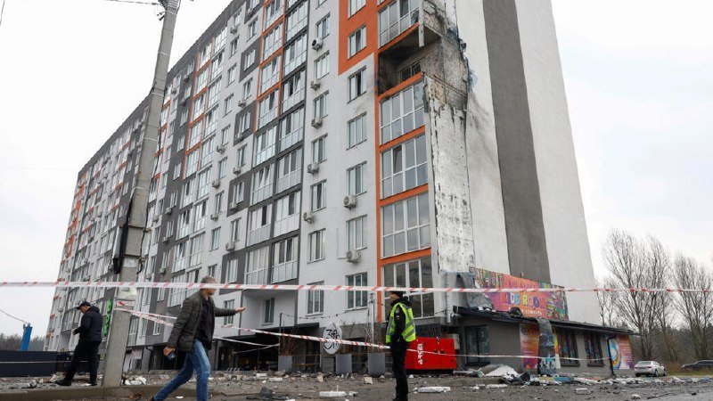 A multi-story residential building shows severe structural damage with a partially collapsed facade, cordoned off by red and white tape while pedestrians and a worker in a high-visibility vest stand nearby amid scattered debris.