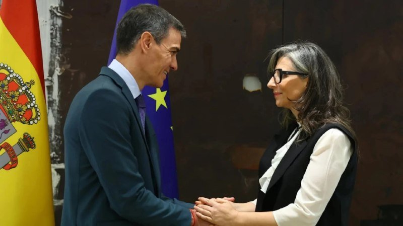 A man in a suit and a woman in a vest shake hands in front of a Spanish flag and an EU flag.