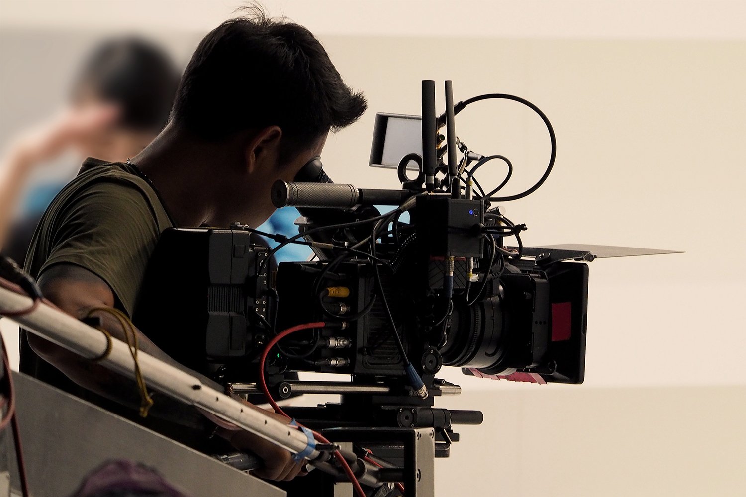 A cameraman in a green shirt looks through the viewfinder of a large professional video camera equipped with antennas and monitors, operating on an outdoor film set.