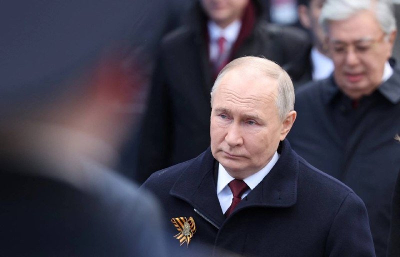 A balding man in a dark coat with a St. George ribbon pin stands among a crowd of people in dark clothing.