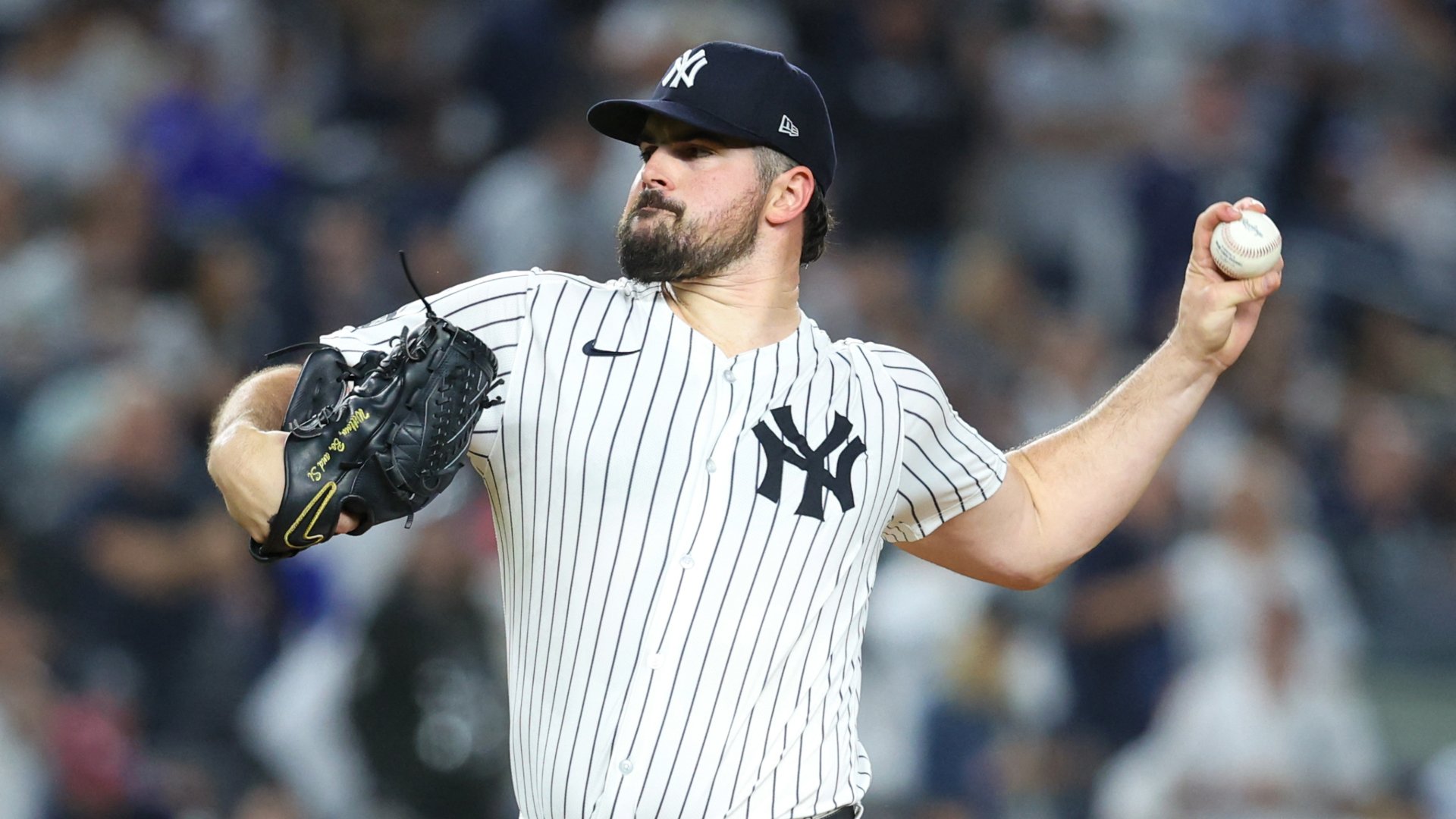 A bearded New York Yankees pitcher in a pinstriped uniform winds up to throw a baseball during a game in front of a blurred crowd.