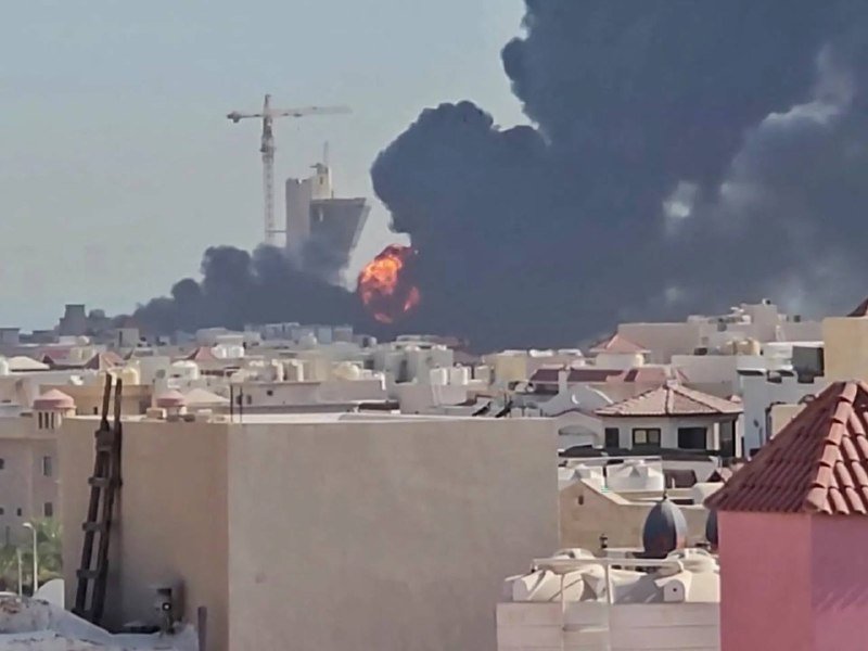 A large fire with a massive plume of thick black smoke rises behind a row of low-rise residential buildings and a construction crane.