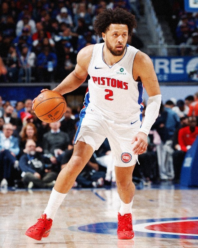 A basketball player wearing a white Pistons jersey with the number 2 dribbles a ball on the court during a game, with spectators visible in the background.