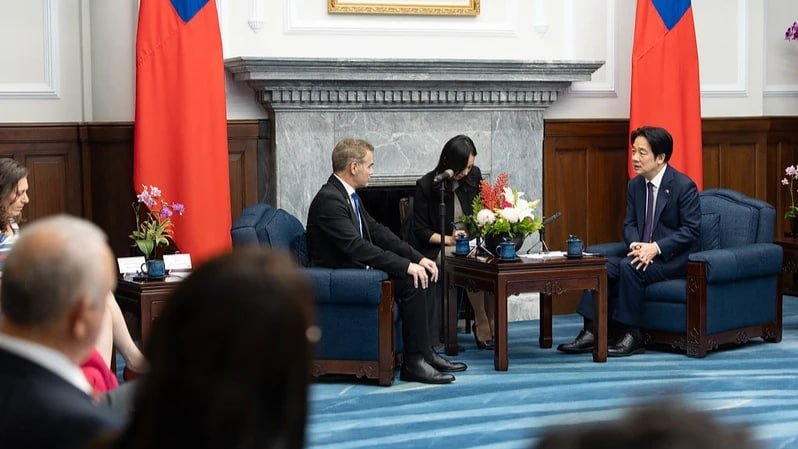Two men in suits sit in blue armchairs facing each other across a small table, with a woman standing between them near a fireplace and two red-and-blue flags.