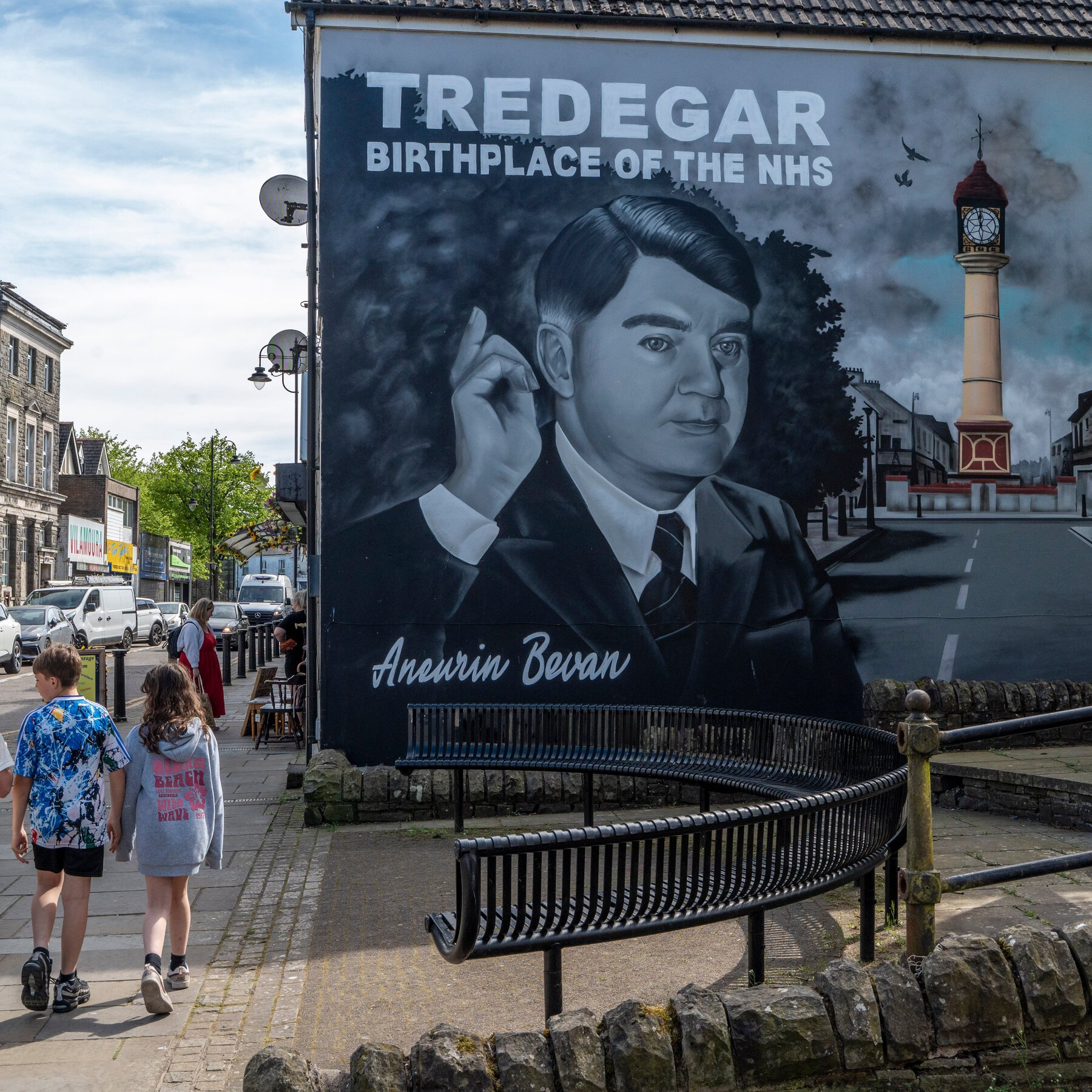 A large mural reading "TREDEGAR BIRTHPLACE OF THE NHS" depicts a man labeled "Aneurin Bevan," with pedestrians walking past on a town sidewalk.