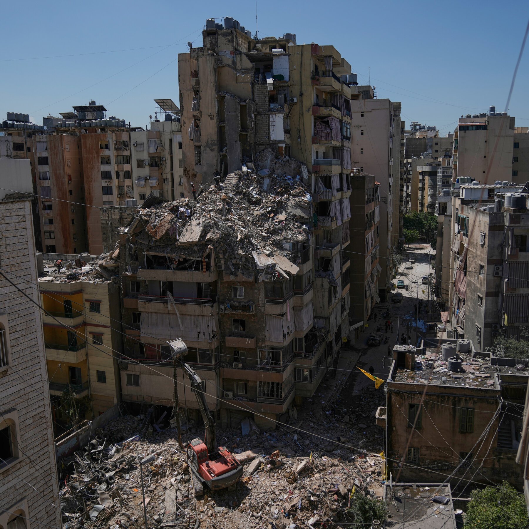 An aerial view shows a heavily damaged multi-story apartment building with a large pile of rubble cascading down its side, as an orange excavator clears debris amid densely packed residential structures under a clear blue sky.