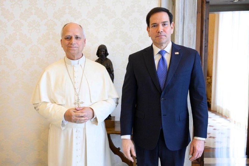 Two men pose side by side in a formal room: one in white papal robes with a cross necklace, the other in a dark suit and blue tie.