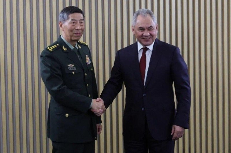 A military officer in a green uniform shakes hands with a man in a dark suit and red tie indoors against a paneled wall.