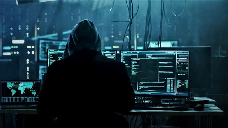 A hooded figure sits before multiple computer monitors displaying code, data graphs, and a world map in a dimly lit, blue-tinted room.