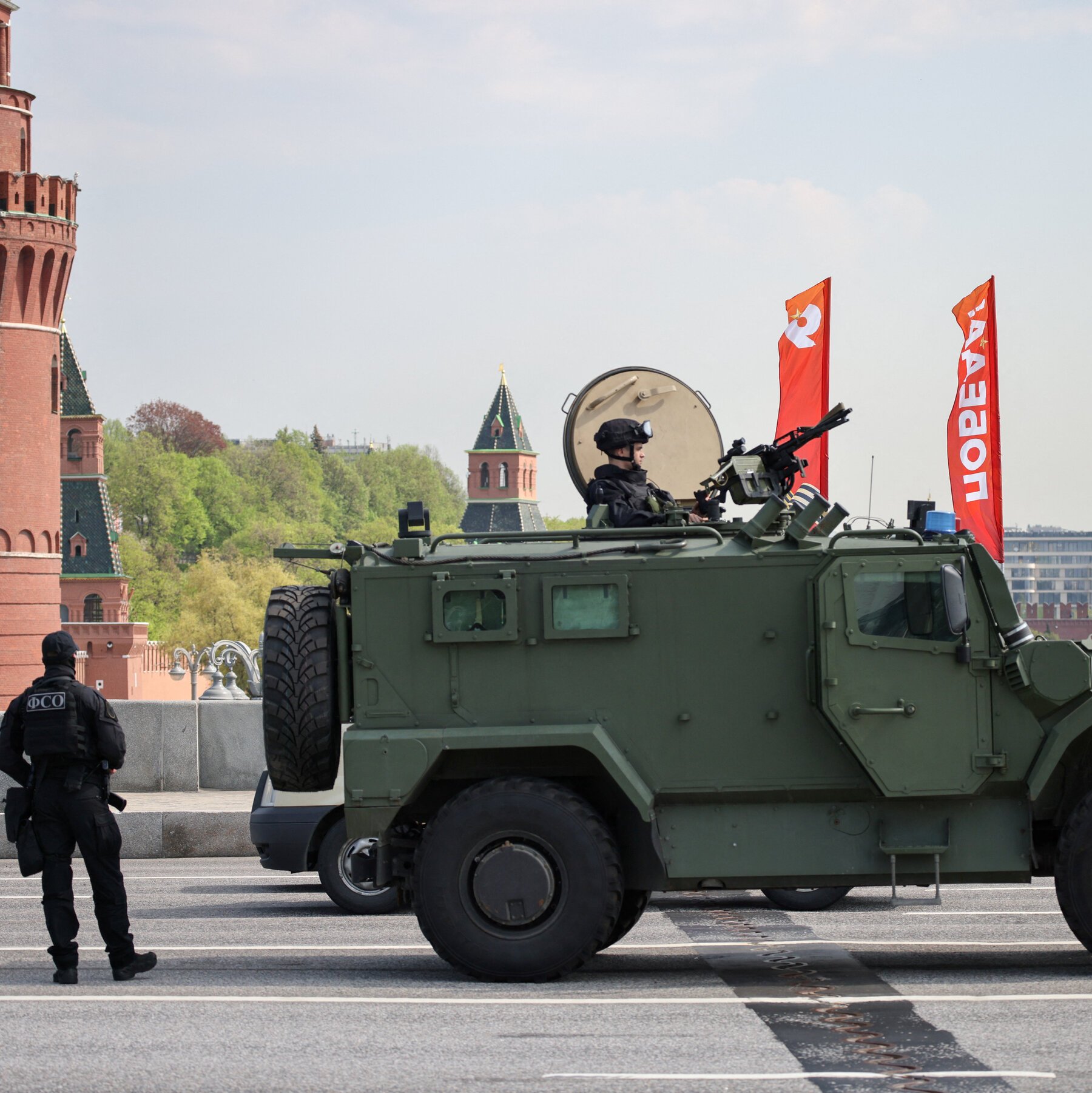 A green armored vehicle with a mounted gunner travels across a paved area, while a uniformed officer with "ФСБ" on his vest stands nearby, with red Kremlin towers and a "9 МАЯ" banner in the background.