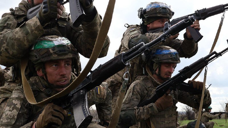 Four soldiers in camouflage uniforms and helmets grip rifles while standing in a grassy field under a cloudy sky, with blue-and-yellow tape visible on their gear.