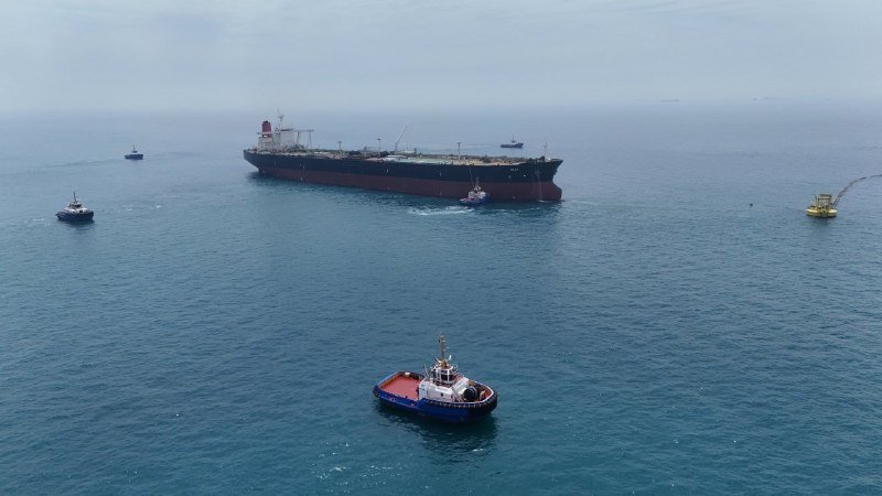 Aerial view of a large oil tanker accompanied by several smaller tugboats and support vessels on calm, open water under a hazy sky.