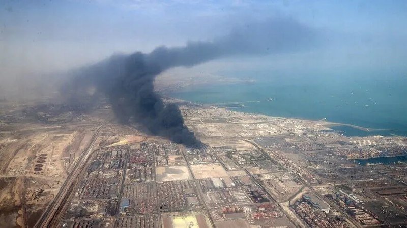 Aerial view of a coastal industrial port area with a large plume of thick black smoke rising from a fire.