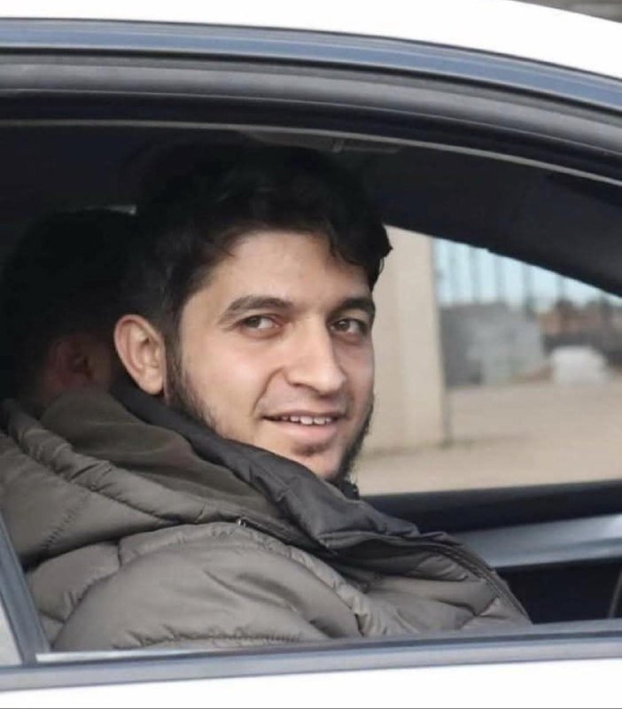 A young man with dark hair and a beard smiles while sitting in the back seat of a car, viewed through the open window.