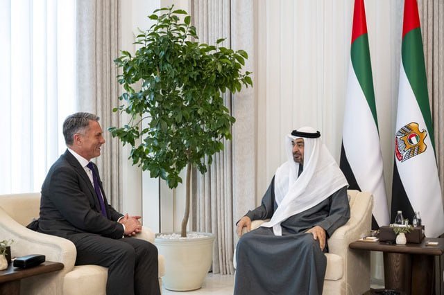 Two men sit facing each other in a formal meeting room, one in a dark Western suit and the other in traditional white Gulf attire, with UAE flags displayed in the background.