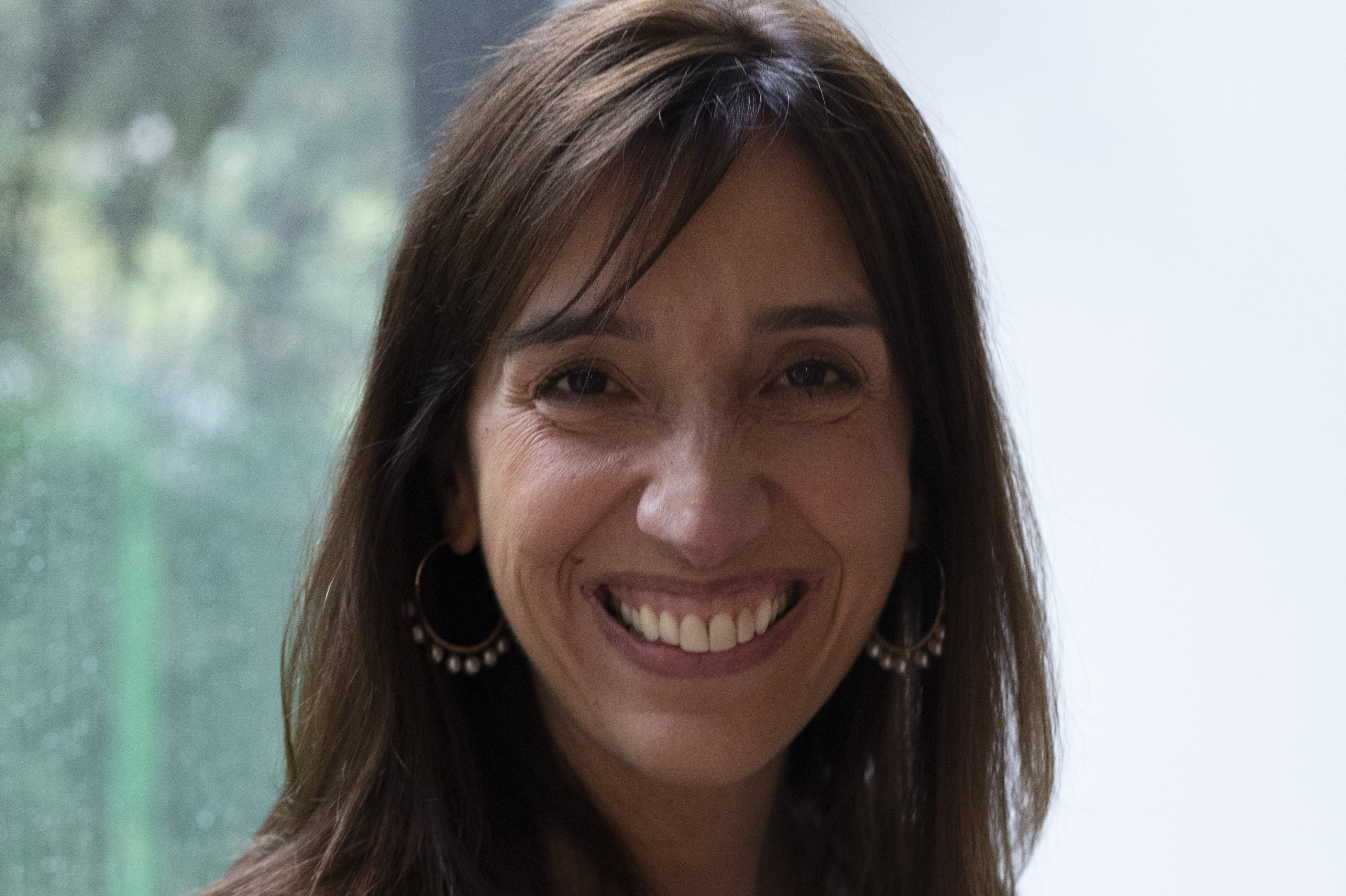 A woman with shoulder-length brown hair and hoop earrings smiles at the camera, with a blurred painting visible in the background.