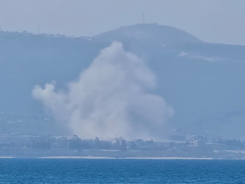 A large plume of white smoke rises from a coastal town across a body of water, with hazy hills and a tower visible in the background.