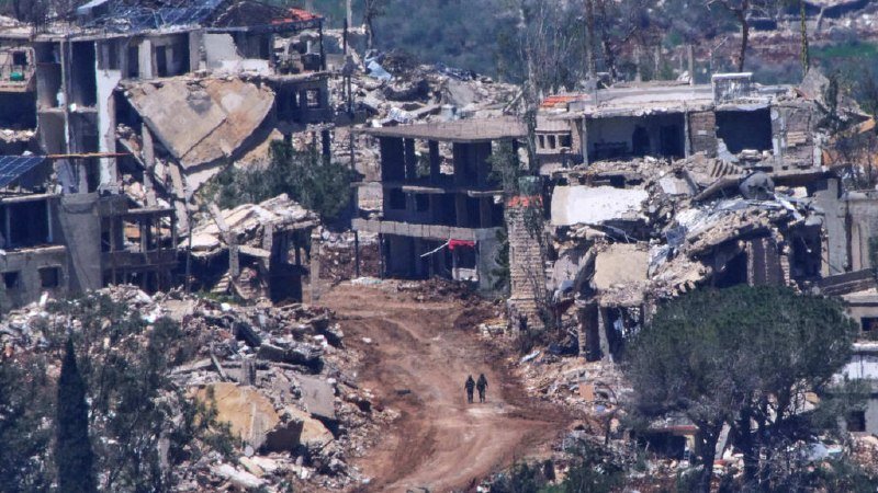 Two small figures walk along a dirt path through extensive rubble and heavily damaged, partially collapsed multi-story buildings.