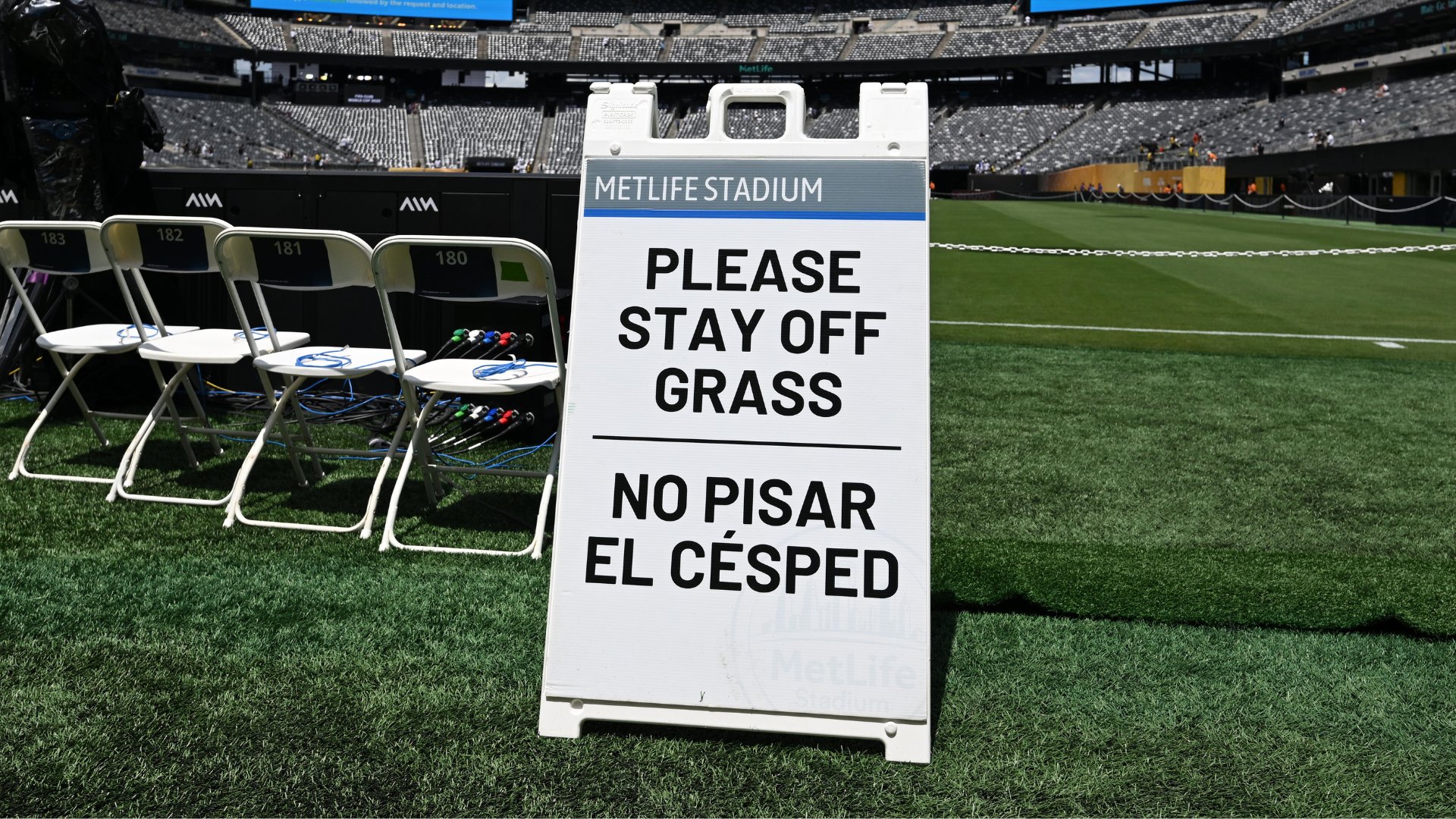 A bilingual "Please Stay Off Grass" sign stands on the field at MetLife Stadium, with numbered folding chairs and stadium seating visible in the background.