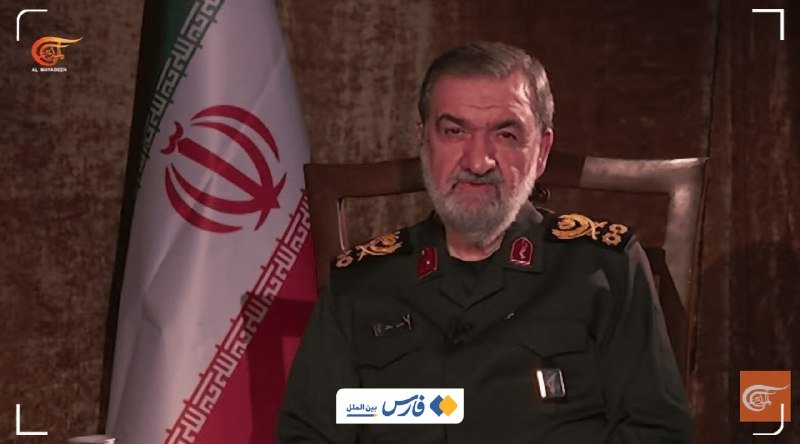 A bearded man in an Iranian military uniform sits seated in front of an Iranian flag, speaking during what appears to be a televised interview.