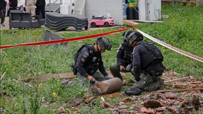 Three uniformed officers in tactical gear kneel in a grassy yard strewn with bricks and rubble, examining a large cylindrical metal object behind red caution tape.