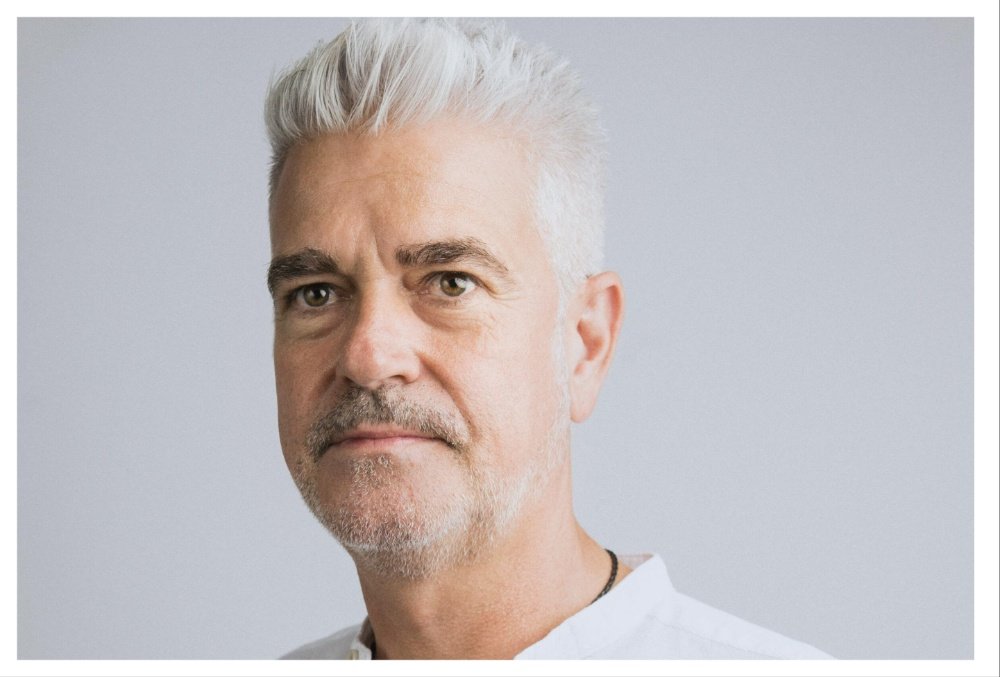 A silver-haired, gray-bearded man poses for a close-up portrait against a plain light gray background, wearing a white top and a black cord necklace.