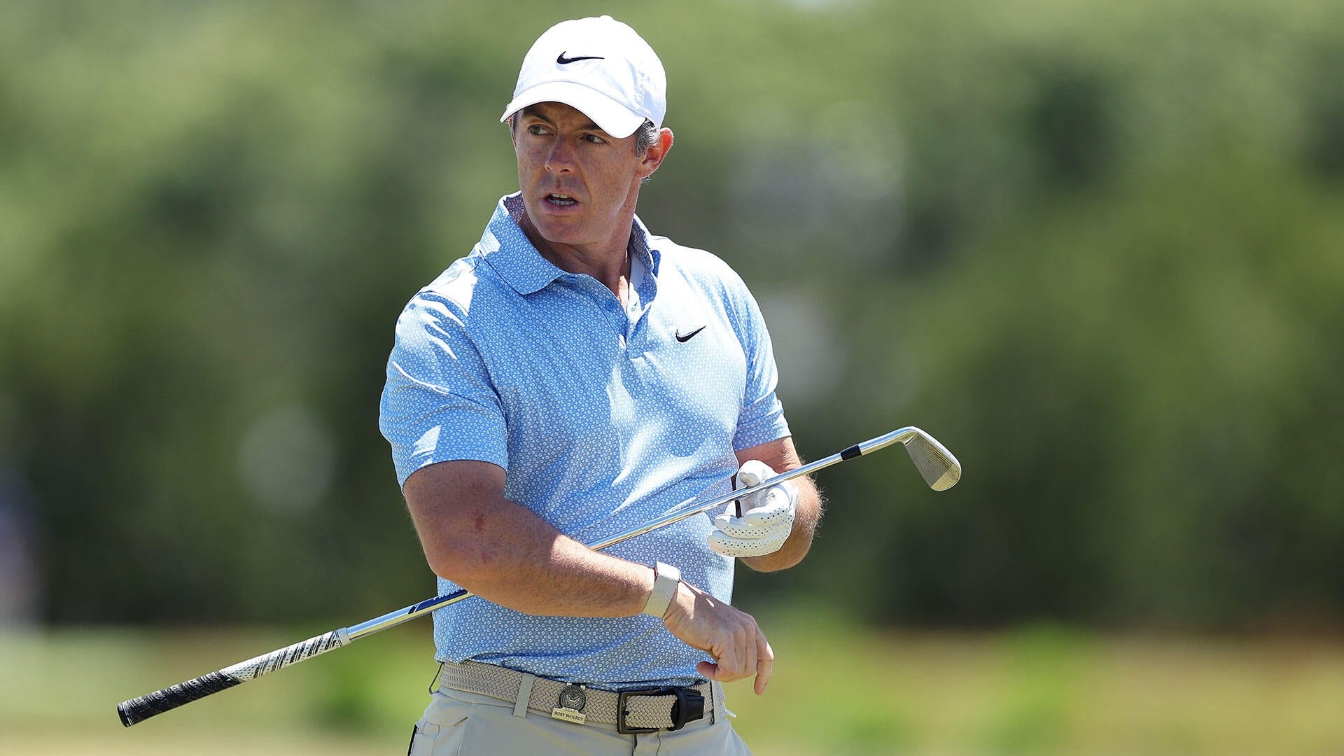A golfer wearing a white Nike cap, light blue patterned polo shirt, and white glove holds an iron club on a sunlit grassy course.