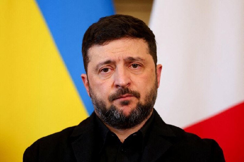 A bearded man in a black shirt and jacket looks to the side, with blue-yellow and white-red flags visible in the background.
