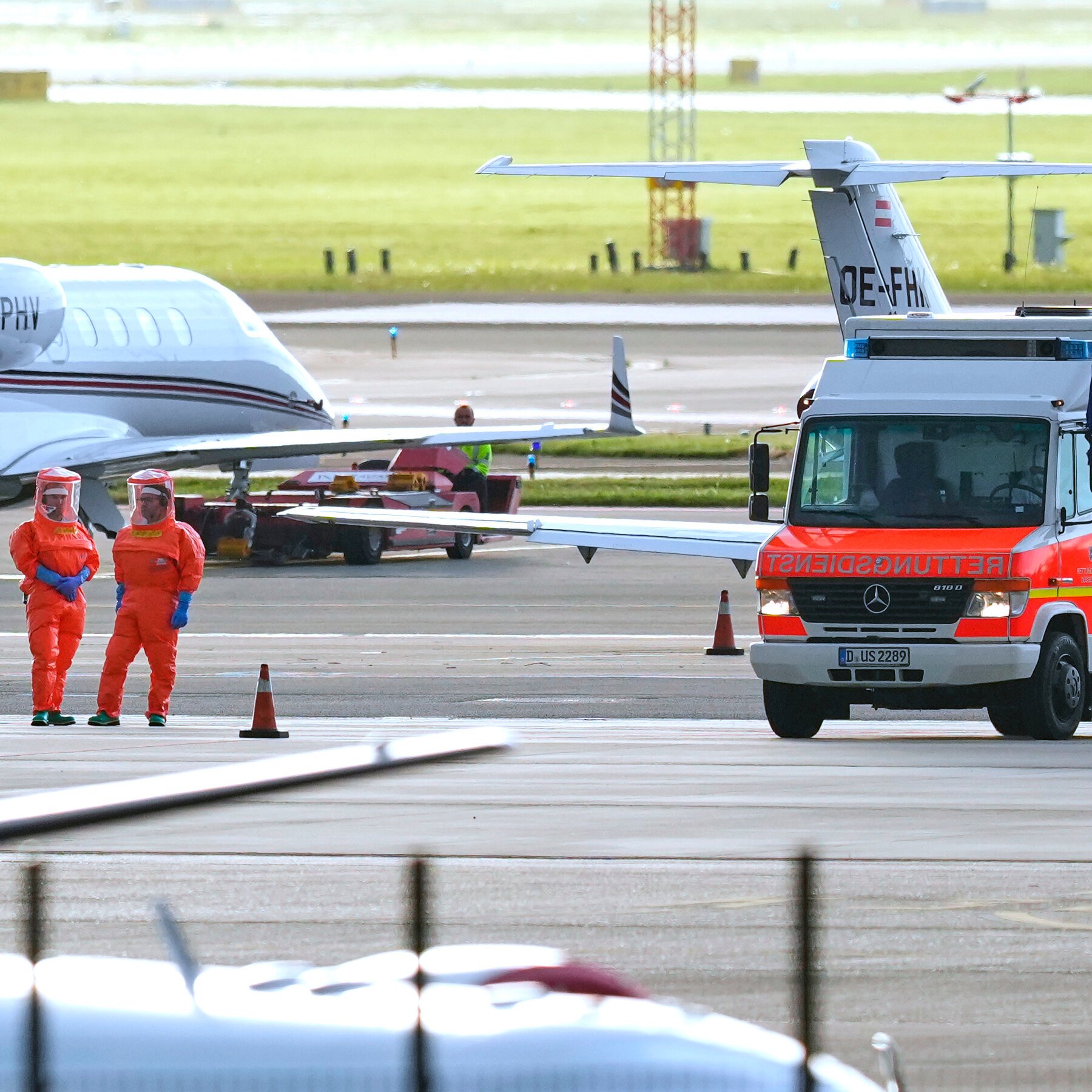 Two personnel in orange hazmat suits stand on an airport tarmac near traffic cones, a red-and-white Rettungsdienst emergency vehicle, and parked private jets.