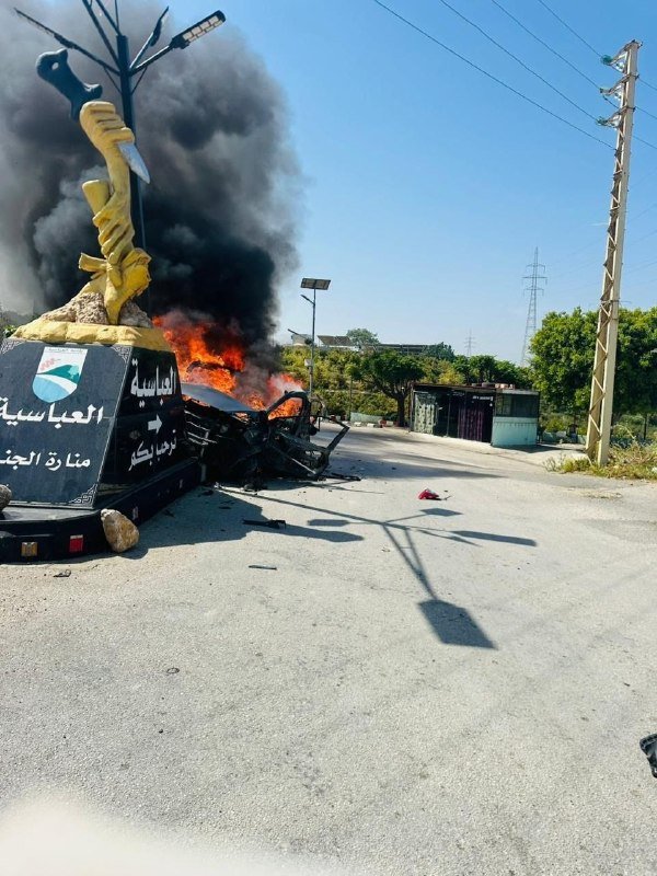 Thick black smoke rises from a burning vehicle beside a yellow monument with Arabic inscriptions on a paved road near power lines.
