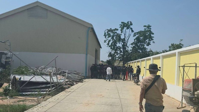 A group of people gathers between a large metal warehouse and a yellow perimeter wall along a paved walkway, with debris piled nearby.